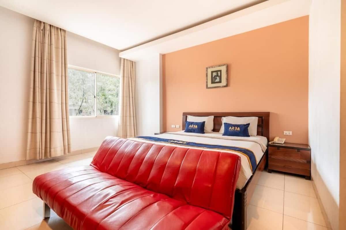 Ein Hotelzimmer mit einem Bett, einer roten Lederbank, einem Fenster mit Vorhängen, einem Nachttisch und einem Wandbild.