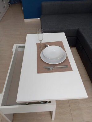 Une table à manger avec une assiette blanche, un verre d’eau et des couverts, disposés sur un set de table marron dans une pièce moderne avec un canapé gris et un mur bleu.