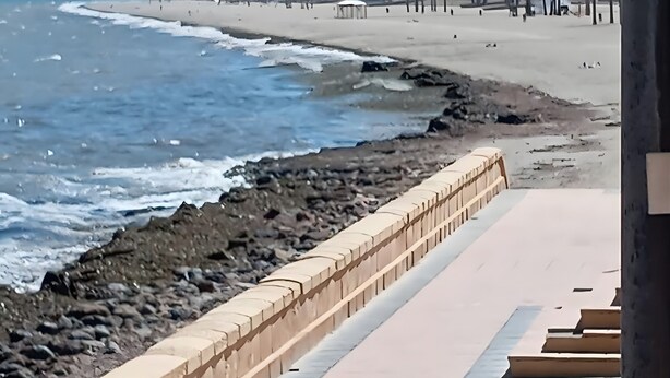 Ein Küstenweg mit Steinmauer, Strand und Gebäuden im Hintergrund.