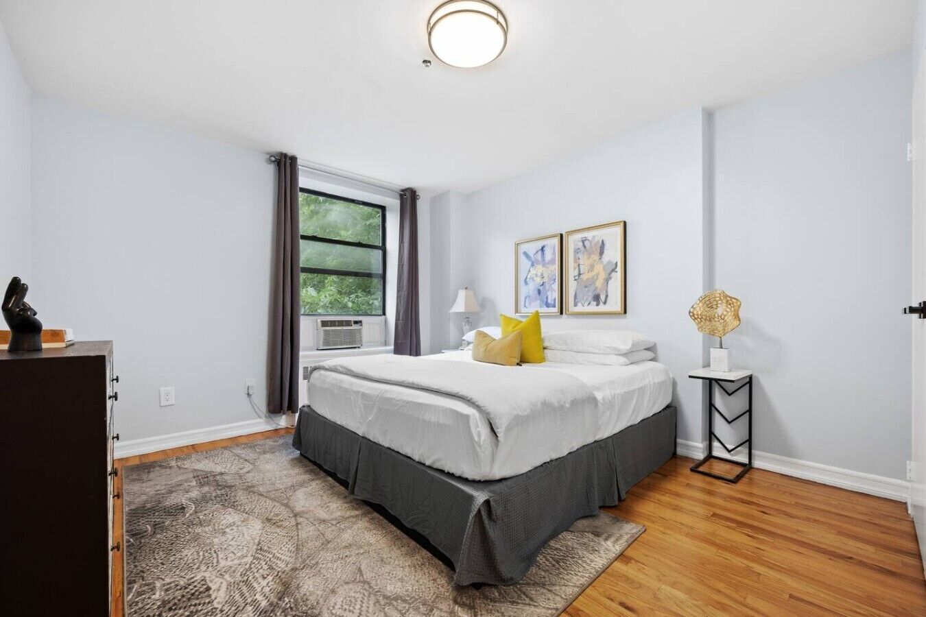 Un dormitorio con cama, mesita de noche, lámpara, ventana con cortinas y una obra de arte en la pared.