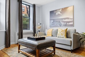 Ein modernes Wohnzimmer mit einem Lederhocker, einem grauen Sofa mit gemusterten Kissen, einem großen gerahmten Kunstwerk an der Wand und Blick durch ein Fenster.