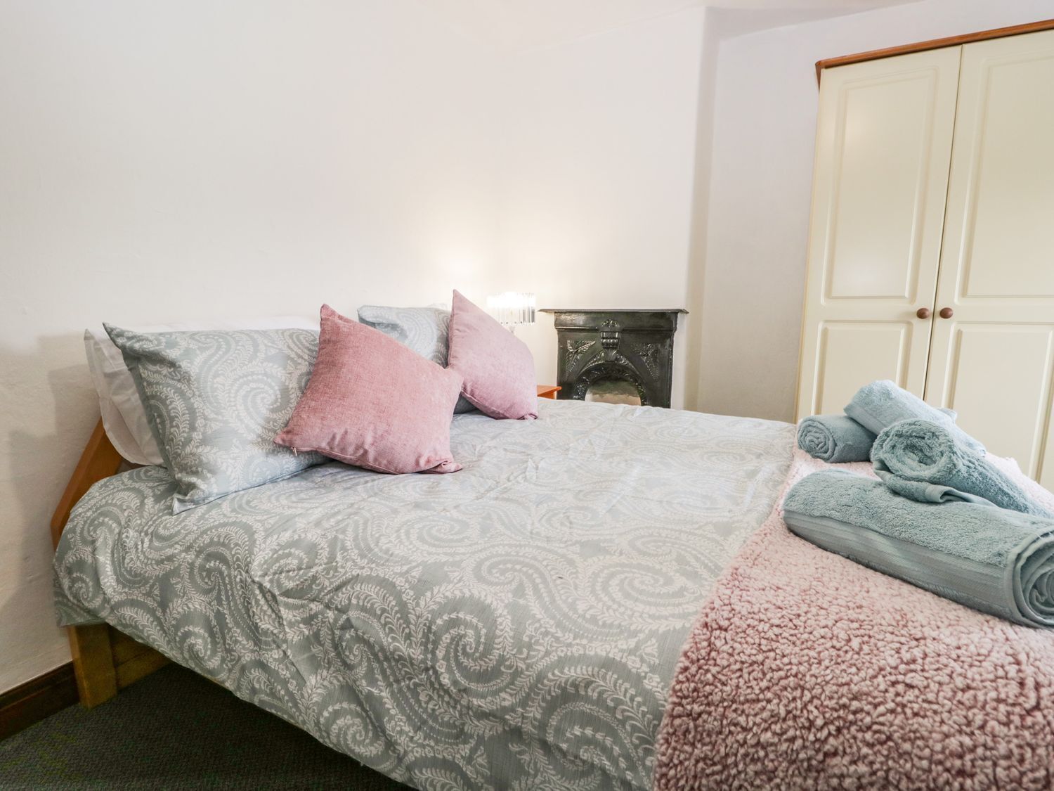 A neatly made bed with patterned bedding, a small bedside table with a lamp, and a wardrobe in a room with plain walls.