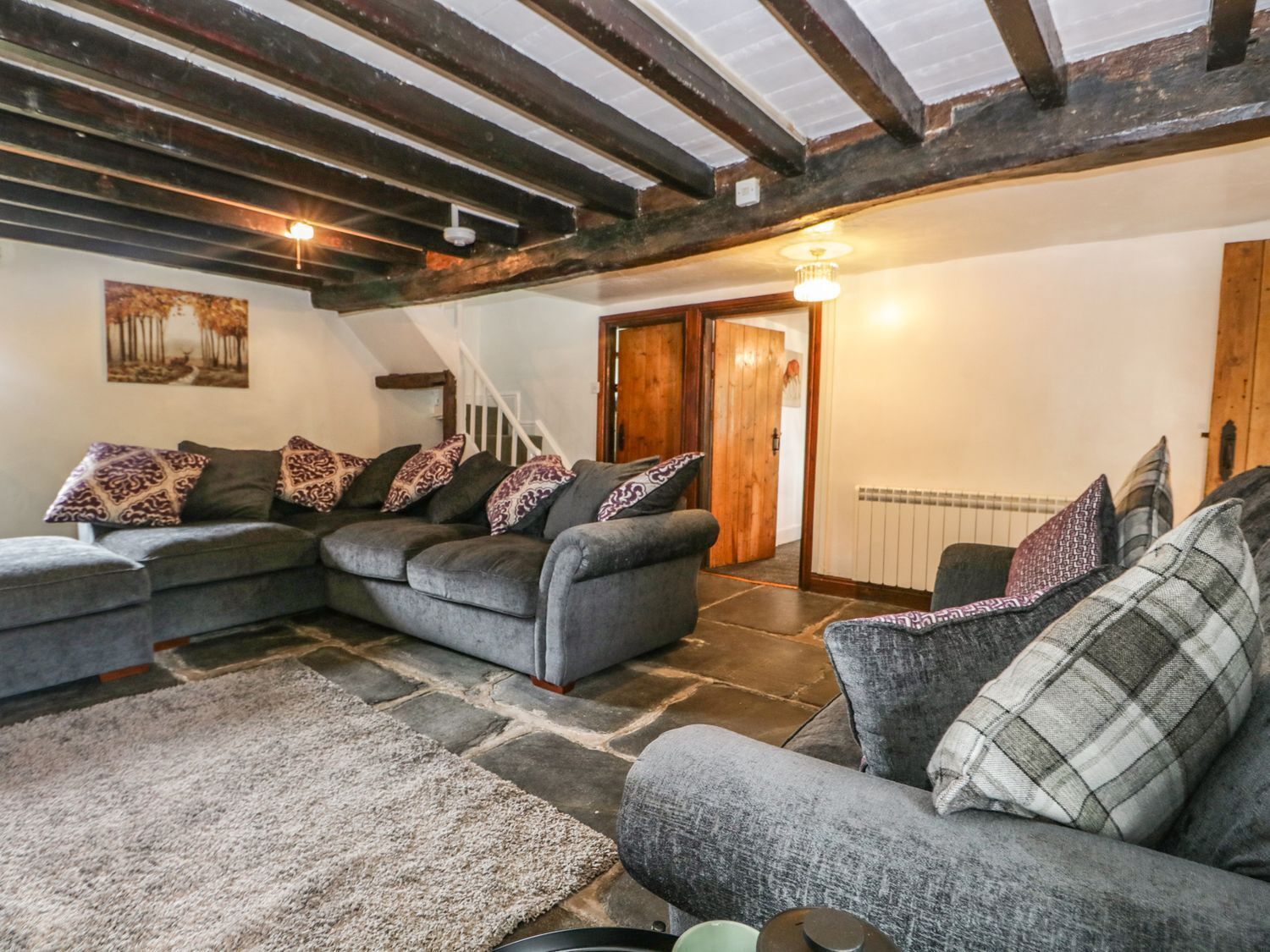 A cozy living room with a sectional sofa, patterned cushions, a stone floor, exposed wooden beams, a staircase, and a radiator.