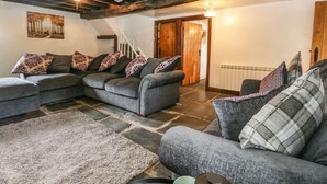 A cozy living room with a sectional sofa, patterned cushions, a stone floor, exposed wooden beams, a staircase, and a radiator.
