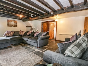 A cozy living room with a sectional sofa, patterned cushions, a stone floor, exposed wooden beams, a staircase, and a radiator.