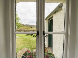 Une fenêtre donnant sur un espace verdoyant, un mur de pierre et un bâtiment à l’extérieur blanc et à la porte verte.