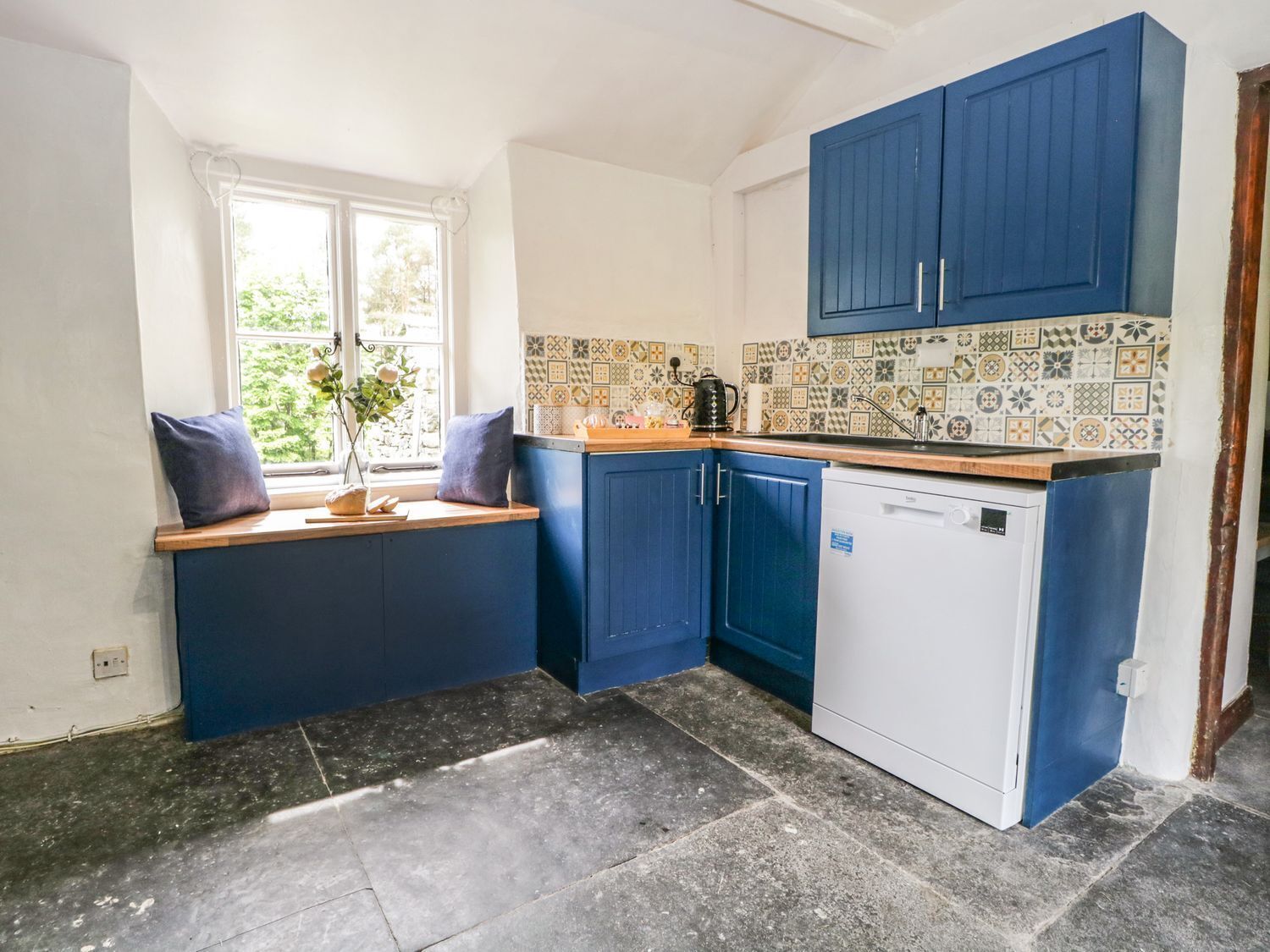 A kitchen with blue cabinets, a white dishwasher, and a window with a view of greenery.