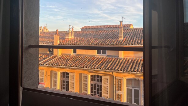 Vista a través de una ventana que muestra un edificio con varios niveles de techo, chimeneas visibles y cielo despejado.