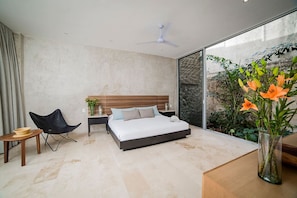 Ein modernes Schlafzimmer mit Bett, Sessel, Nachttisch und einem großen Fenster mit Blick auf Grünflächen.