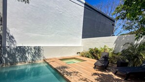 Een modern buitenzwembad met een houten terras, twee ligbedden en een kleine jacuzzi.