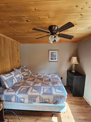 A bedroom with a wooden ceiling, a ceiling fan, a bed with a quilt, a dresser, and a framed picture on the wall.
