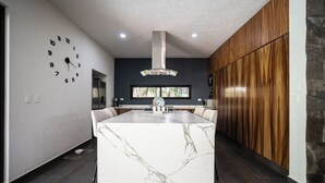 A modern kitchen with a marble island, wooden cabinets, and a clock on the wall. - Luxury Pool Home | Sleeps 8 | TV Room + 4 Full Baths (Playa del Carmen)