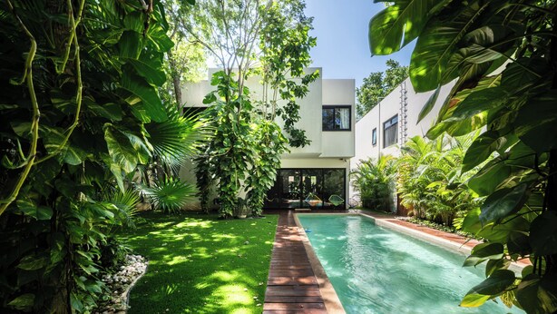 A modern pool area with a wooden deck, surrounded by lush greenery and a building with large windows. - Luxury Pool Home | Sleeps 8 | TV Room + 4 Full Baths (Playa del Carmen)