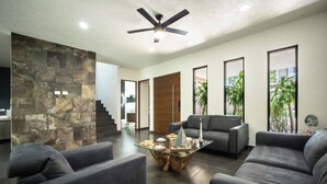 A modern living room with a stone accent wall, a glass coffee table, grey sofas, and a ceiling fan. - Luxury Pool Home | Sleeps 8 | TV Room + 4 Full Baths (Playa del Carmen)