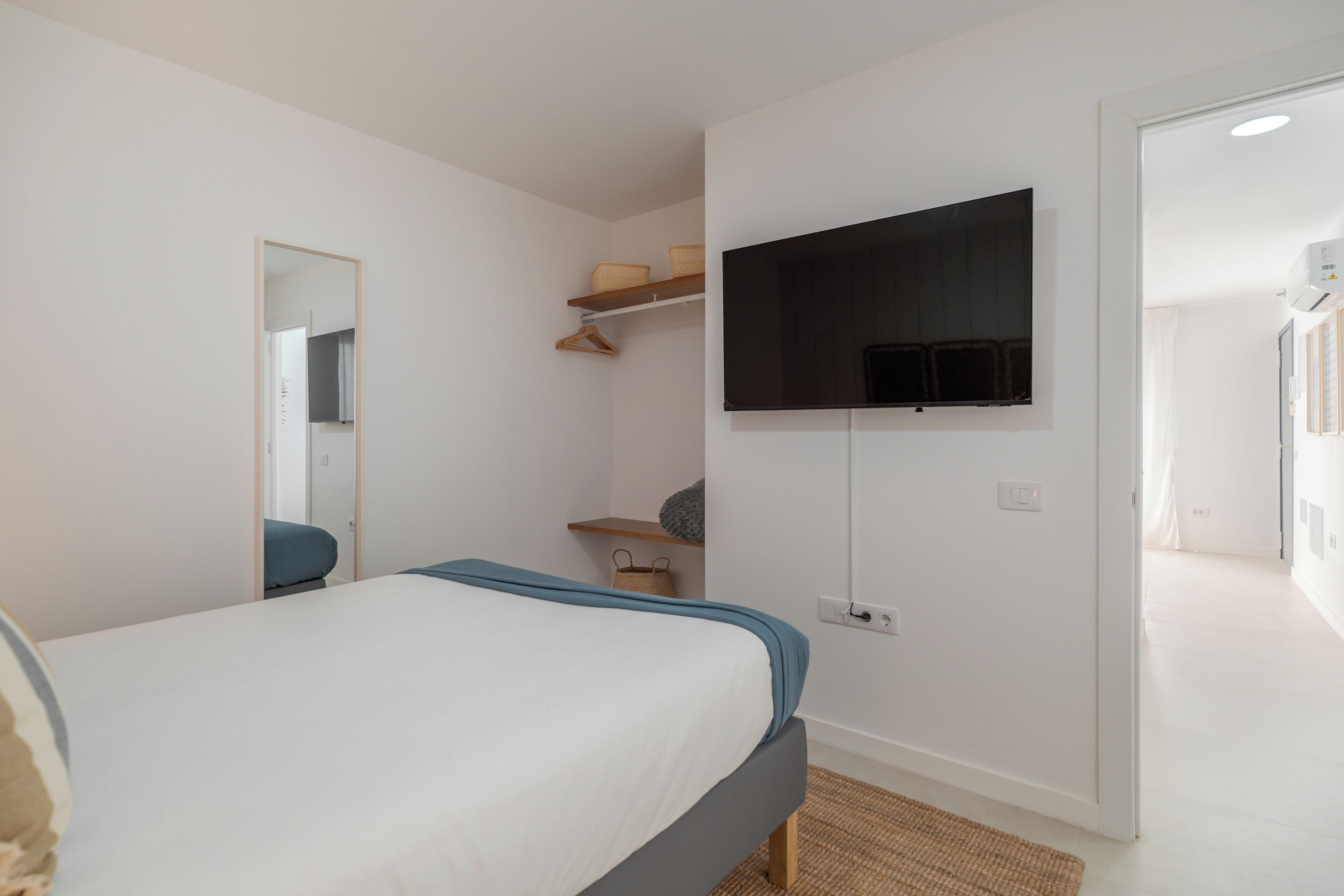 Un dormitorio con una cama, televisor montado, un espejo y una puerta que da a otra habitación.