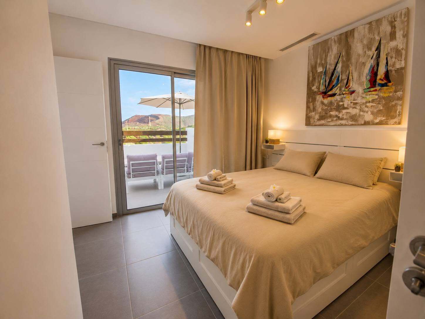 A hotel room with a bed, a painting, a balcony with chairs, and a view of the landscape.