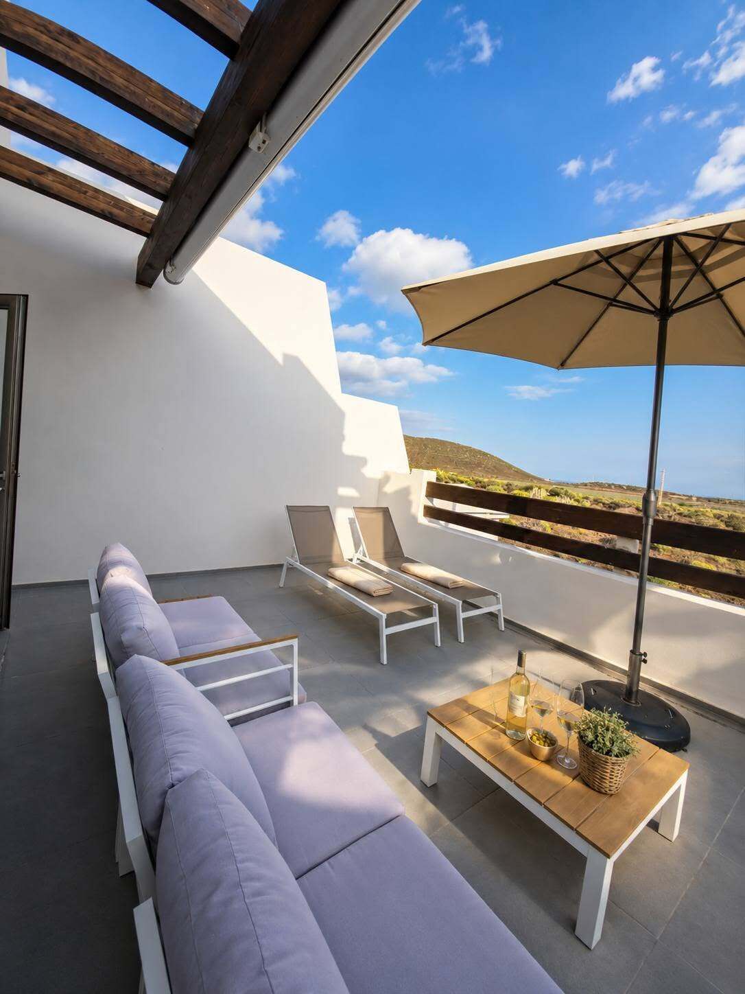 Une terrasse sur le toit avec un canapé blanc, deux fauteuils de relaxation, une table en bois avec une bouteille et des verres, et un grand parasol.
