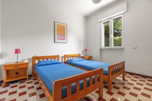 Een slaapkamer met twee houten bedden, een nachtkastje, een lamp en een raam met uitzicht op groen.