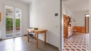 Ein Zimmer mit Holztisch, einem Stuhl, einer Schiebetür, die auf einen Balkon führt, sowie einer Küchenzeile mit Holzausstattung und gefliestem Boden.