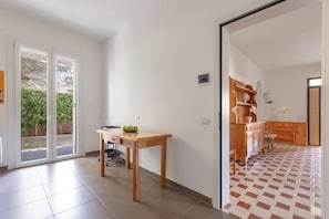 Ein Zimmer mit Holztisch, einem Stuhl, einer Schiebetür, die auf einen Balkon führt, sowie einer Küchenzeile mit Holzausstattung und gefliestem Boden.