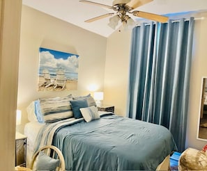 Una camera da letto con un letto, un ventilatore a soffitto, un comodino, una lampada, un quadro appeso al muro e una finestra con tende.