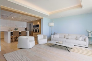 Ein modernes Wohnzimmer mit einer Couch, einem Couchtisch und Sesseln. Eine Küche mit weißen Schränken sowie ein Essbereich mit Tisch und Stühlen sind sichtbar.