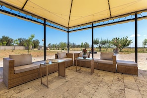 Un espace extĂ©rieur abritĂ©, amĂ©nagĂ© avec des meubles en osier et une table, entourĂ© dâun ciel dĂ©gagĂ© et dâarbres Ă lâhorizon.