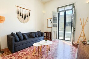 Ein Wohnzimmer mit einer dunklen Couch, einem runden weißen Tisch, einer Wandweberei aus Makramee und einem Holzeasel.