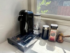 Een koffiezetapparaat, een glazen kan, een metalen beker, een kruidenpot en een koffiemolen op een aanrecht bij een raam.