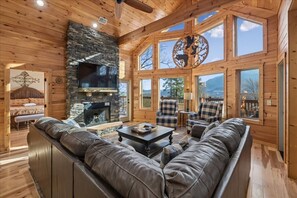 Ein Blockhaus-Interieur mit Kamin aus Stein, einer Ledersofa und großen Fenstern mit Blick auf die Berge.