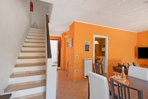 Una escalera conduce a una sala de estar con paredes anaranjadas, una mesa de comedor con sillas y una zona de cocina al fondo.