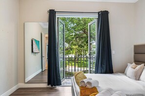 Ein Schlafzimmer mit Bett, einem fenster mit Vorhängen, Blick auf Grünfläche draußen und einer Tür zu einem anderen Raum.