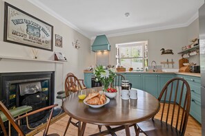 A dining area with a table set for breakfast, a fireplace, teal cabinetry, and a window with a view. - Smithy Cottage -  Beauly , Scottish Highlands, Scotland (Beauly)