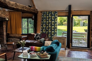 A cozy living room with a leather sofa, a wooden coffee table with books, a brick fireplace, and a view of the garden through large windows. - SPACIOUS COTTAGE BETWEEN LAND AND SEA - Sleeps 9, 5 bedrooms, 2 shower rooms, 3 toilets, ping-pong table (Veulettes-sur-Mer)