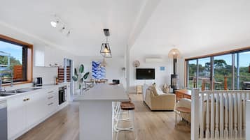A modern kitchen with a central island, bar stools, a sofa, and a TV in a living area with a large window.