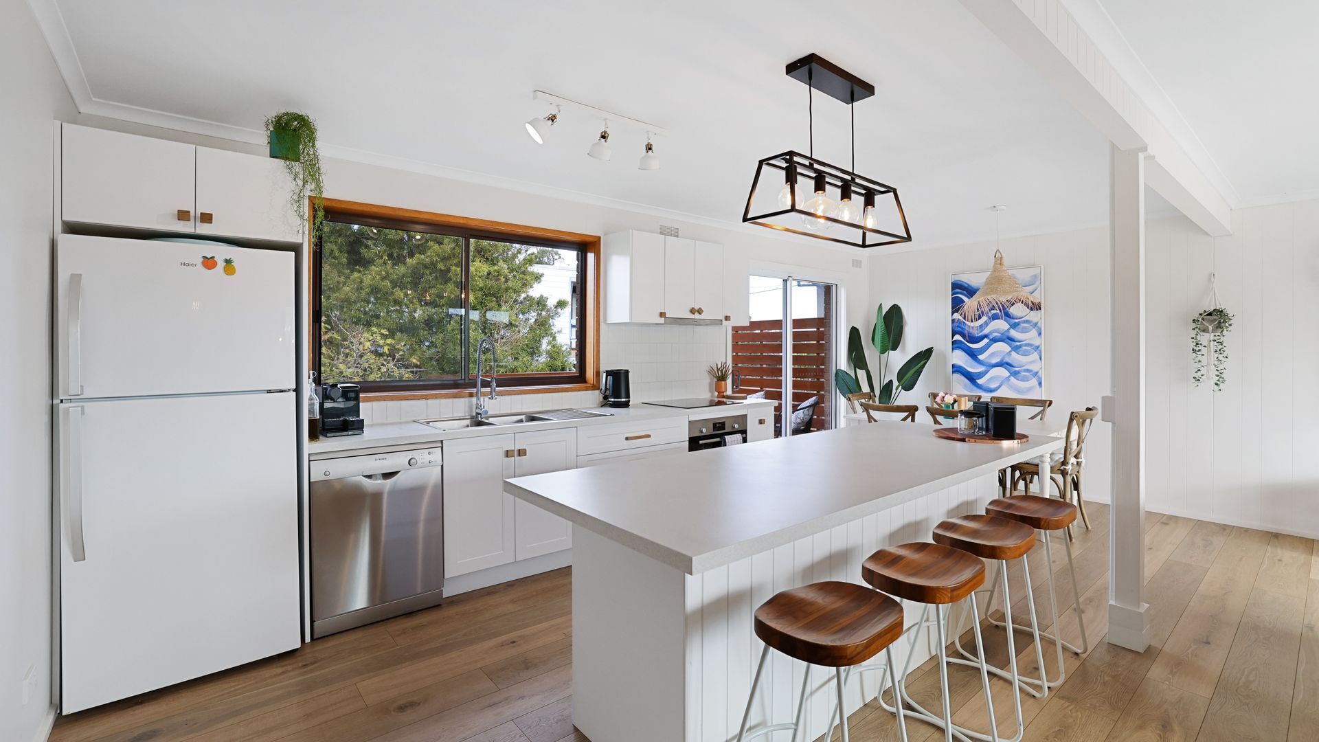 A modern kitchen with white cabinetry, a large island with stools, a built-in dishwasher, and a window with a view of trees.