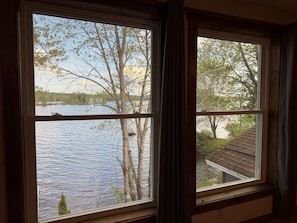 A room with a large window overlooking a lake, a wooden window frame, and a view of trees and a building across the water.