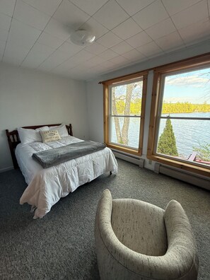 Ein Schlafzimmer mit Bett, Sessel, Fenster mit Seeblick und einer Deckenleuchte.