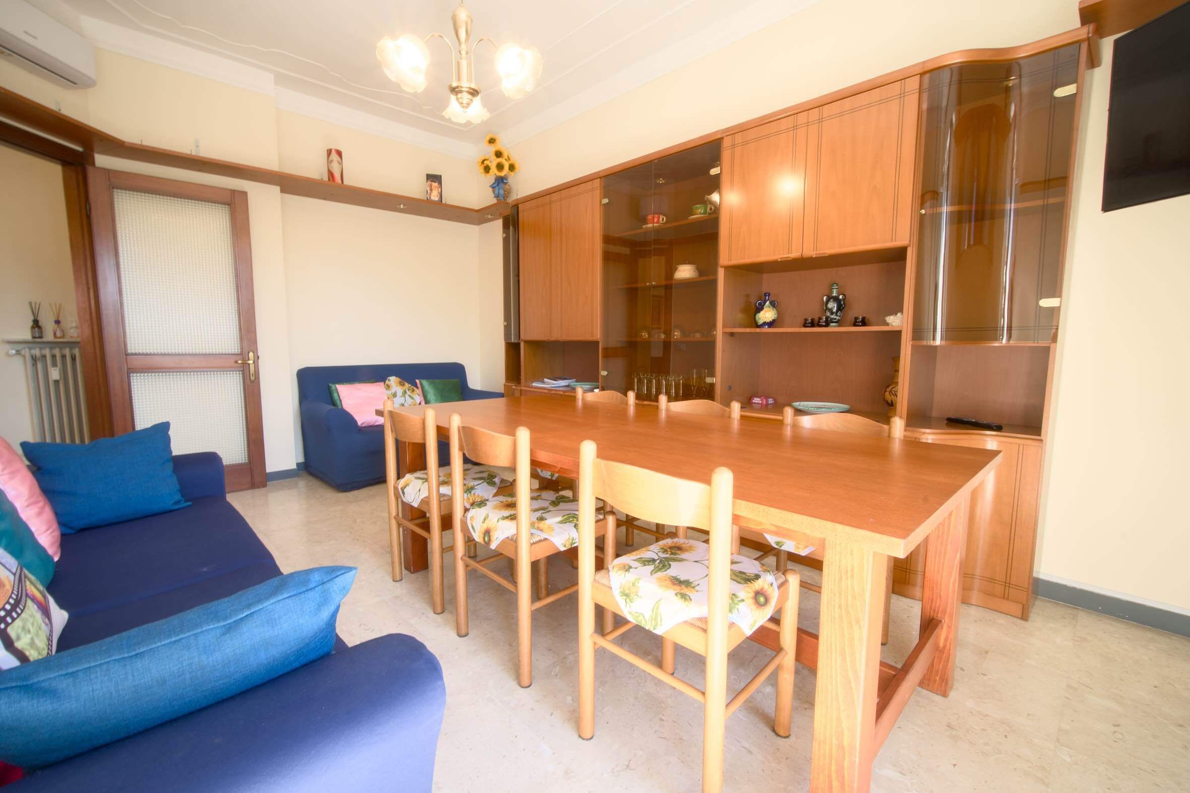 A dining area with a wooden table and chairs, a sofa, and a kitchen area with cabinets and a refrigerator.