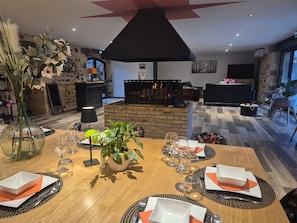 Un coin repas agrémenté d’une cheminée en briques, une table dressée pour quatre personnes et une vue sur le salon, avec son canapé et sa télévision.