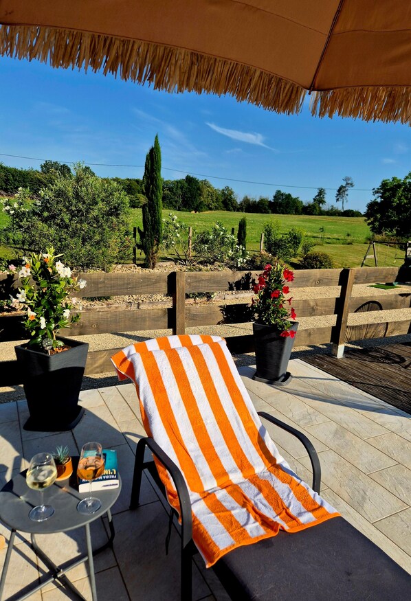 A lounge chair with a striped towel, a small table with a glass of wine and a book, and potted plants.