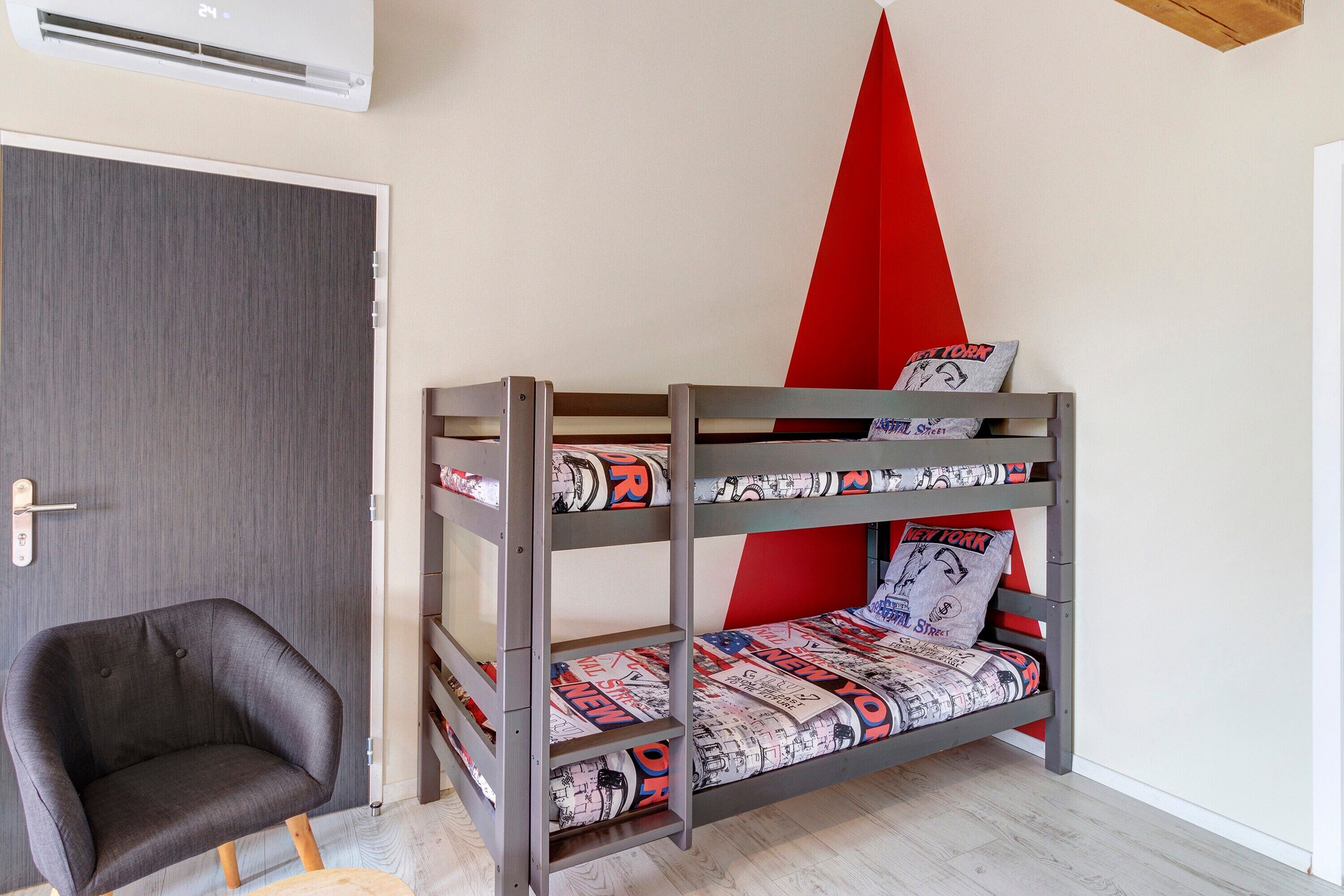 Una cama litera con ropa de cama estampada, un sillón gris y un adorno triangular rojo en la pared.