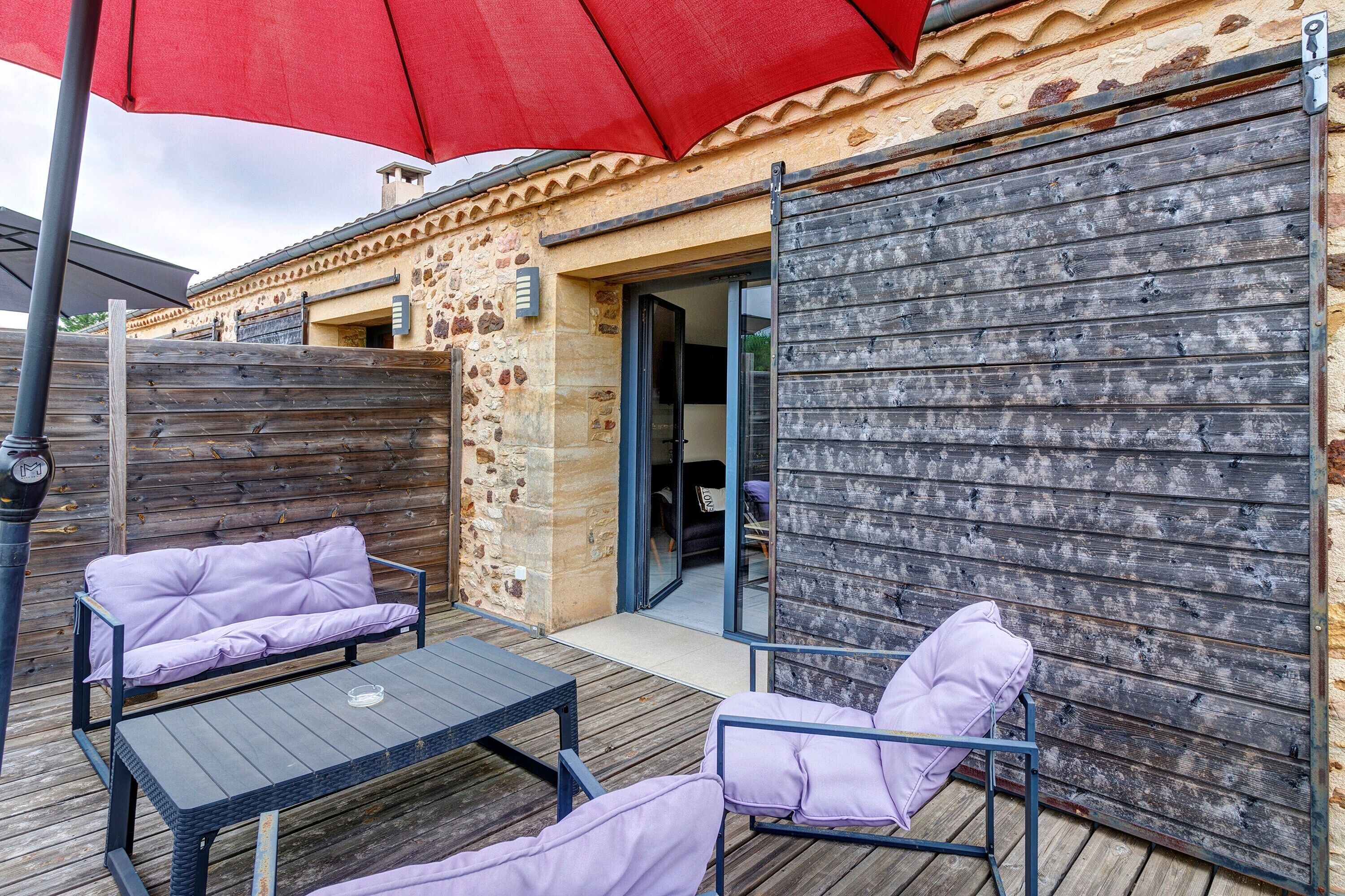 Une terrasse extérieure avec une plateforme en bois, deux fauteuils rembourrés, une table et un parasol rouge.