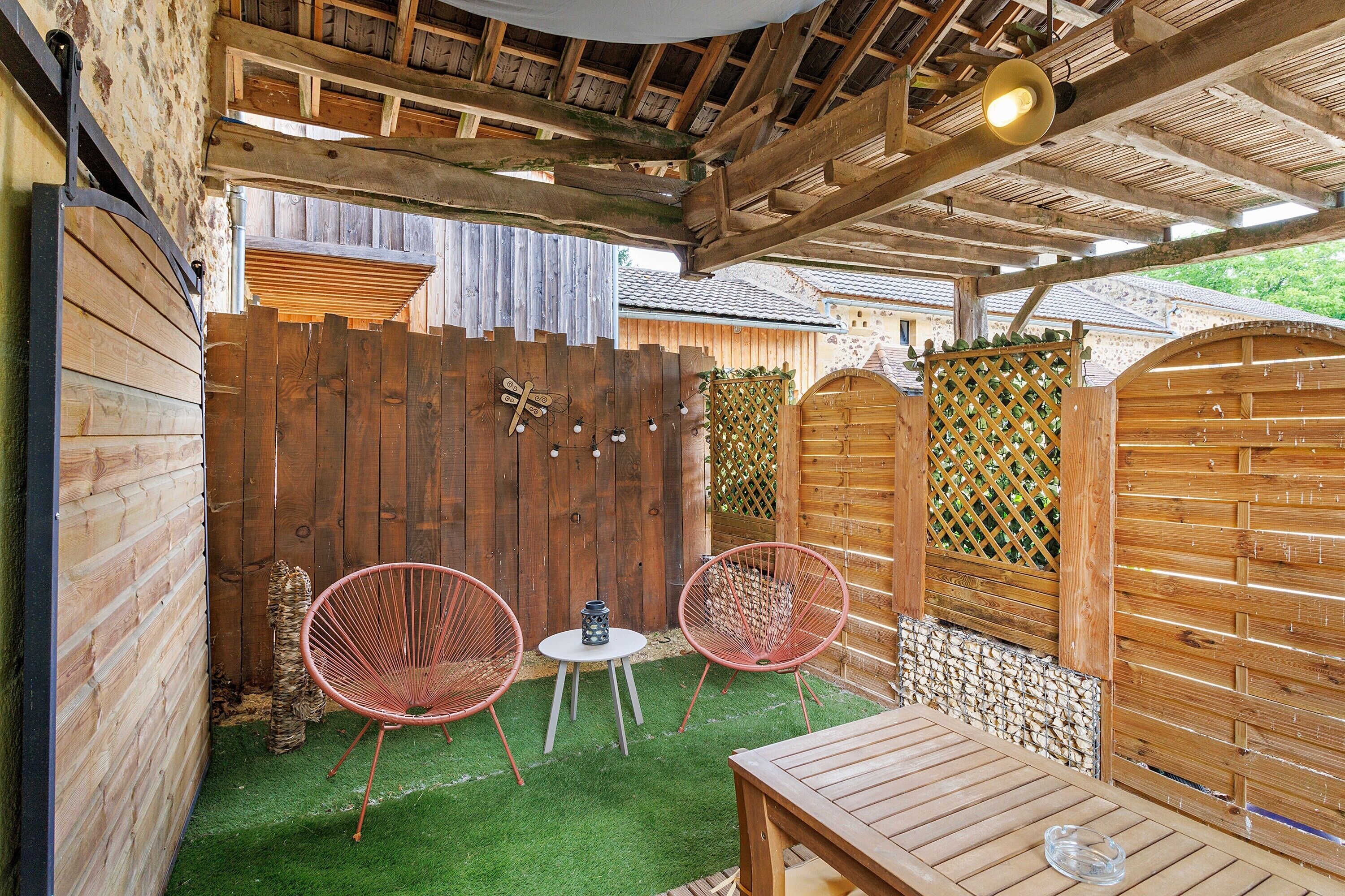 Een houten terras met twee rode draadstoelen, een klein rond tafeltje en een houten tafel.