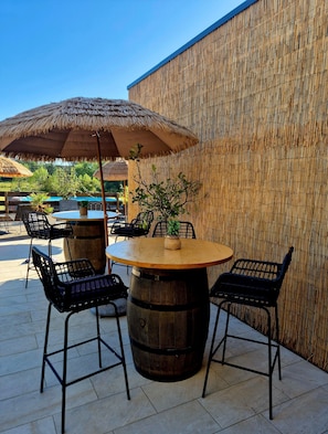 Un espace extérieur aménagé avec une table ronde en bois, des chaises en métal noir et un parasol en chaume.