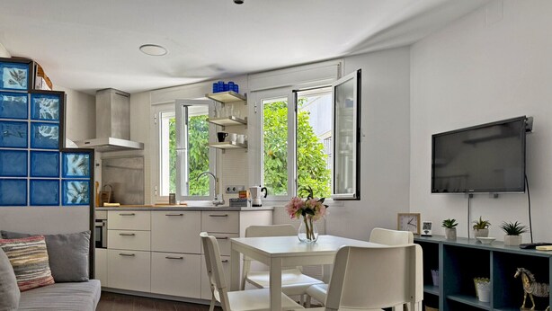 Une cuisine moderne avec des meubles blancs, une table à manger avec des chaises, un téléviseur fixé au mur et une fenêtre donnant sur un paysage verdoyant.