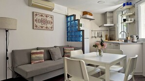 Une cuisine moderne avec des meubles blancs, un coin repas avec une table et des chaises blanches, un canapé gris agrémenté de coussins à motifs et une fenêtre à cadre bleu.