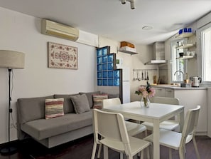 Eine moderne Küche mit weißen Schränken, ein Essbereich mit weißem Tisch und Stühlen, ein graues Sofa mit gemusterten Kissen und ein Fenster mit blauem Rahmen.