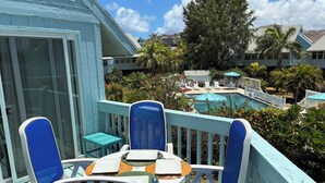 Varanda com mesa e cadeiras, com vista para área de piscina com guarda-sóis e espreguiçadeiras.