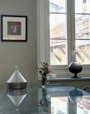 Una habitación con un cuadro en la pared, un florero con flores sobre una mesa y una ventana que ofrece vistas a edificios.